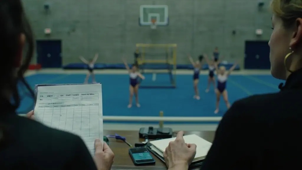 Parent seated in gym bleachers holding a printed session rotation sheet while gymnasts in matching leotards warm up on the floor mat